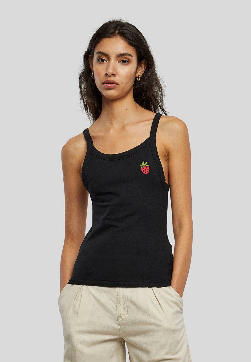 Jeune femme aux longs cheveux foncés porte un débardeur noir avec un petit motif fraise rouge et un pantalon beige clair, mains dans les poches.