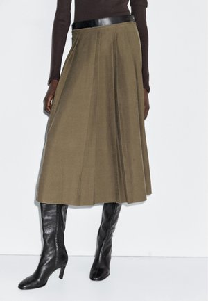 Femme portant une jupe midi plissée vert olive avec une ceinture noire, associée à des bottes en cuir noires montantes à talons.