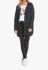 Lang mørkeblå strikket cardigan med åben front, tekstureret stof og ribbede manchetter, parret med en hvid grafisk t-shirt og sorte slim fit jeans.