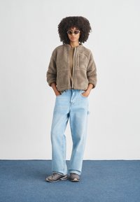 Žena s kučeravými vlasmi, ktorá nosí slnečné okuliare, hnedú fleecovú bundu, svetlomodré široké rifle a sivé tenisky, stojaca s rukami v vreckách.