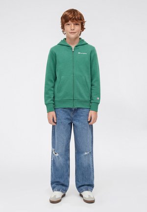 Groene hoodie met ritssluiting en zakken, met een logo op de borst. Gecombineerd met lichtblauwe, beschadigde wijde broekspijpen en witte sneakers.