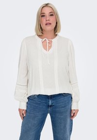 White blouse with a V-neck and tie detail, featuring textured polka dots, vertical pintucks, and gathered sleeves. Paired with blue jeans.
