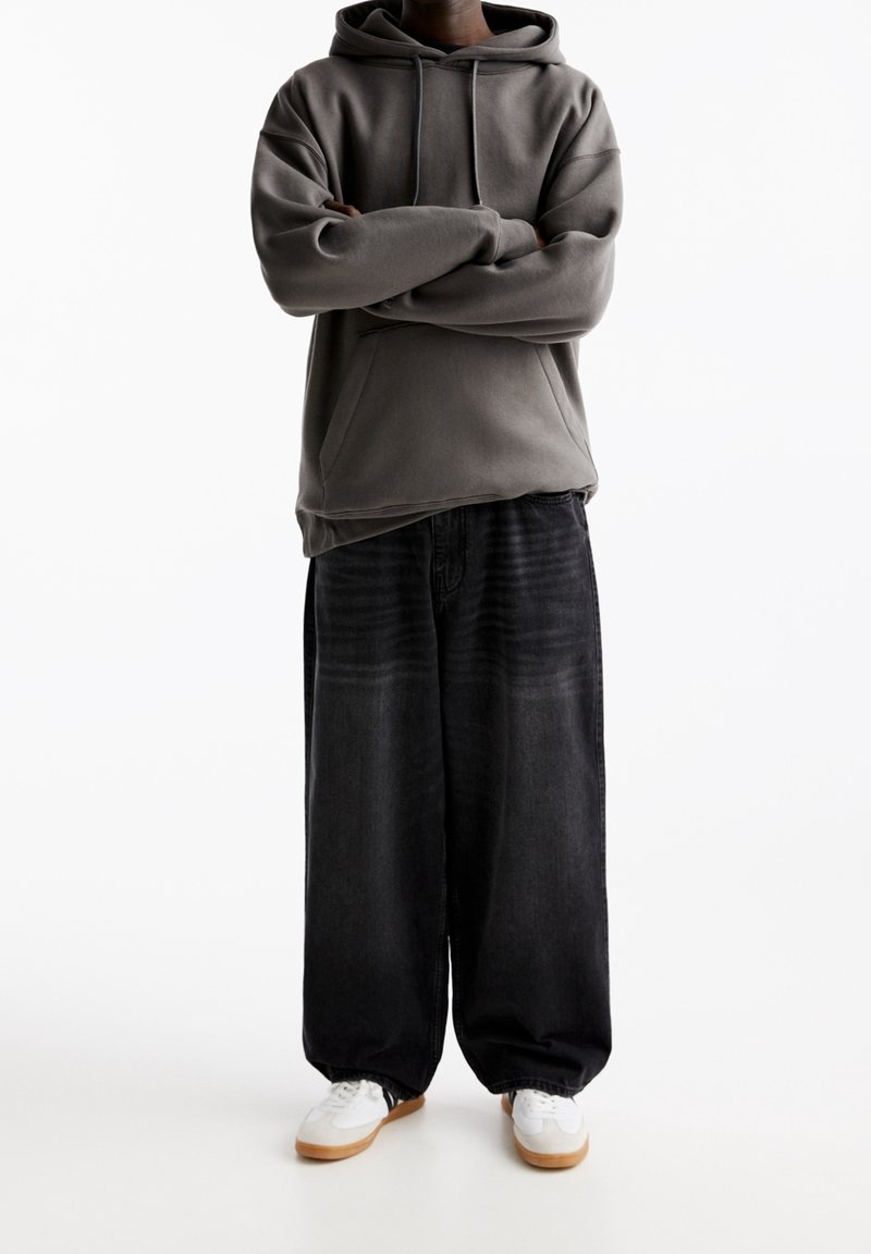 Personne portant un sweat à capuche gris foncé, un jean noir large et des baskets blanches, debout les bras croisés sur un fond blanc.