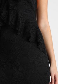 Vestido negro de encaje con un patrón texturizado y detalles con volante en el escote. La tela parece suave e intrincada.