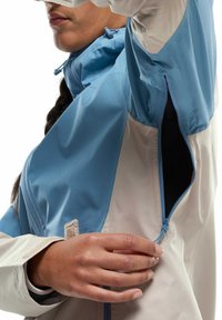 Leichte Jacke mit einem zweifarbigen Design, bestehend aus einem blauen oberen Teil und einem cremefarbenen unteren Teil. Enthält eine mit Reißverschluss versehene Tasche und eine verstellbare Kapuze.