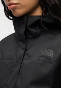 Μαύρο αδιάβροχο μπουφάν με ψηλό γιακά. Διαθέτει ματ λογότυπο The North Face στο στήθος. Ομαλή υφή και μινιμαλιστικός σχεδιασμός.
