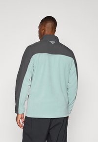 Chaqueta de forro polar con un cuerpo de color turquesa claro y sección superior gris oscuro. Cuello alto con un pequeño detalle del logo en la parte posterior.