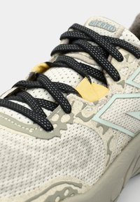 Lys beige snøresko med mesh-overdel, sorte snører, gule detaljer og grønn logo. Teksturerte mønstre på siden.
