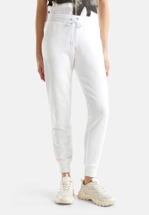 Nedre del af en person, der bærer hvide joggers med snor og beige chunky sneakers, stående mod en ensfarvet lys baggrund.