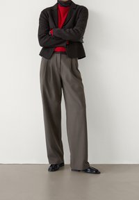 Tummanruskea bleiseri punaisen poolopaidan päällä, harmaat korkeat pussihousut, joissa on laskokset, ja mustat loaferit. Sileä kangas ja istuva malli.