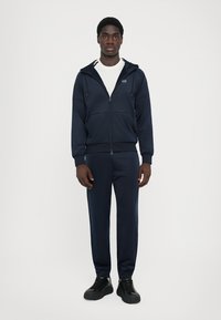 Survêtement de sport marine en tissu léger. Comprend une veste zippée avec poches avant et un pantalon assorti avec des poignets élastiques.