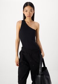Svart randig top med en axel som matchas med svarta joggers med subtila detaljer. Modellen håller en svart tote-väska.