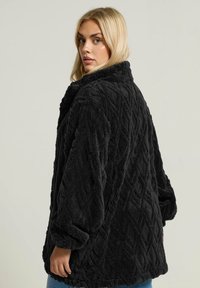 Mustan, suurikokoinen tekoturkisjacketti, jossa on timanttikuosi, pitkät hihat ja pehmeä, teksturoitu pinta. Ominaisuuksiin kuuluu leveä kaulus.