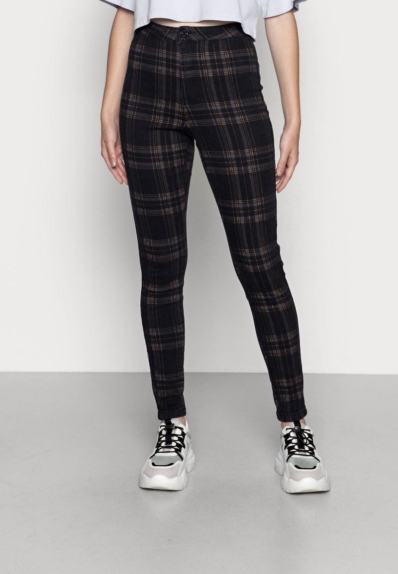 Femme portant un pantalon ajusté à carreaux noirs et marron et des baskets blanches épaisses, debout sur un sol gris devant un mur blanc.