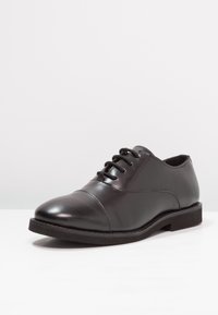 Black leather oxford shoe with laces, rounded toe, and low heel on white surface and light gray background.