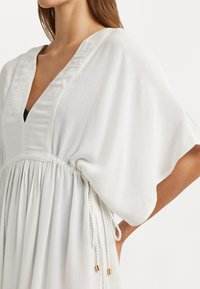 Vestido blanco con un profundo escote en V, mangas cortas y anchas, y una cintura elástica con lazos ajustables. Tela ligera y fluida con una textura suave.