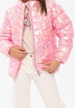 Personne portant une doudoune rose brillante sur un t-shirt graphique beige clair et un pantalon blanc, mains ajustant la fermeture éclair et la poche de la doudoune.