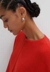 Pull en tricot rouge avec un col rond, associé à des boucles d'oreilles à clou sphériques en argent. Texture lisse et design simple.