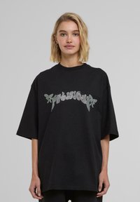 T-shirt nera oversize in cotone, con una stampa lucida di strass che rappresenta un testo stilizzato sul petto. Maniche corte e scollatura rotonda.