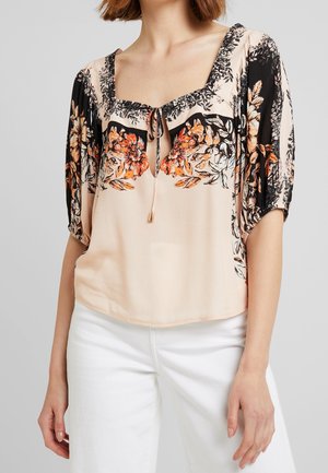 Kobieta ubrana w jasnoróżową bluzkę z czarno-pomarańczowymi kwiatowymi wzorami, z wiązaniem przy dekolcie i bufkami na rękawach, w połączeniu z białymi spodniami.