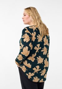 Maglietta a maniche lunghe verde scuro con un motivo floreale beige. Tessuto morbido con una vestibilità comoda e orlo arrotondato. Sono visibili pantaloni neri.