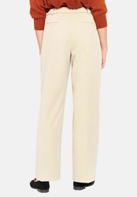 Beige Hose mit geradem Bein, elastischem Bund, Seitentaschen und Nahtdetails auf der Rückseite. Glatte Stoffstruktur, minimalistische Stil.