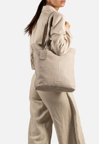 Borsa a spalla in pelle beige con doppi manici, caratterizzata da una sottile etichetta con logo e un design semplice. Texture liscia con una forma strutturata.