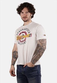 T-shirt in cotone bianco con una grafica di un aereo giallo all'interno di un design circolare rosso, con la scritta "Aeronautica Militare". Maniche corte.