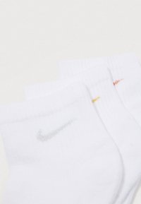 Três pares de meias brancas de tornozelo empilhadas, cada uma com um pequeno logotipo da Nike em prata, amarelo e laranja perto do punho.