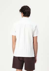Camisa blanca de manga corta con un patrón texturizado y un pliegue en la parte posterior, combinada con pantalones cortos marrones. La camisa presenta un cuello clásico y un corte relajado.