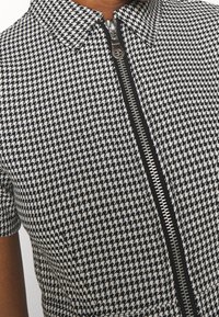 Musta-valkoinen koiranhampaakuviollinen paita, jossa on vetoketjukiinnitys ja terävä kaulus, korostaen teksturoitua kangasta ja lyhyitä hihoja.