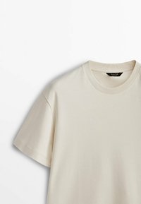 Crèmekleurig T-shirt met korte mouwen en een ronde hals, met een zwart merkembleem aan de binnenkant van de kraag, afgebeeld op een witte achtergrond.