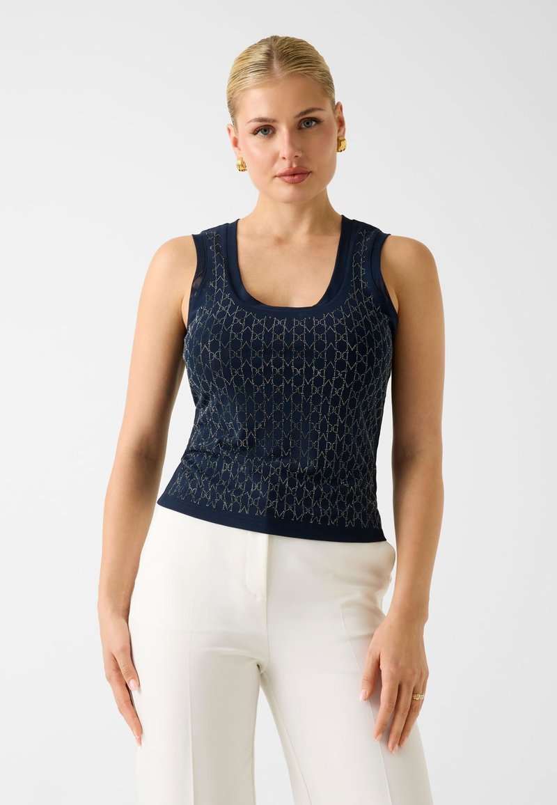 Femme aux cheveux blonds portant un haut sans manches bleu marine avec un motif argenté et un pantalon blanc, debout devant un fond uni clair.