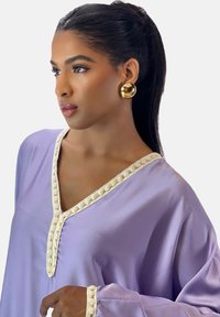 Lavendelkleurige satijnen top met decoratieve crèmekleurige bies langs de halslijn en mouwen. Model draagt gouden cirkelvormige oorbellen en heeft het haar naar achteren gebonden.