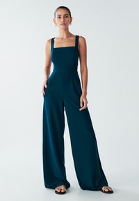 Zelenomodra obleka s širokimi hlačnicami, kvadratnim izrezom in debelimi naramnicami. Izdelana je iz gladkega blaga, ima sprednje šive in je kombinirana z ravnimi sandali.