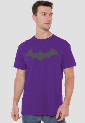 Man wearing a purple t-shirt with a large black Batman logo, looking to the side against a plain light background.