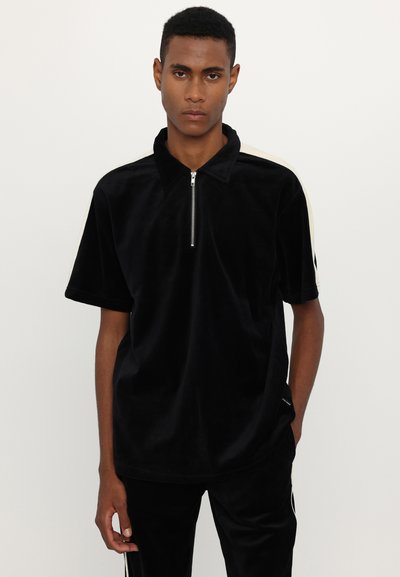 Polo shirt en velours noir avec un col à demi-zip et des manches courtes. Présente des rayures crème contrastantes sur les épaules et une coupe décontractée.