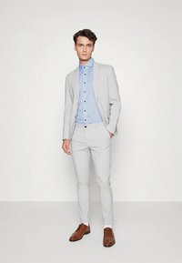 Hellgrauer Anzug mit passender Jacke und Hose. Das blau gestreifte Hemd hat dunkle Knöpfe. Braune Lederschuhe vervollständigen das Outfit.