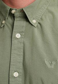 Camicia button-up verde chiaro, con colletto a punta, bottoni in plastica bianchi e piccolo logo ricamato sul petto. Tessuto liscio.