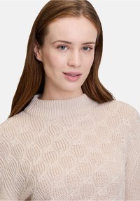 Hellbeige gestrickter Pullover mit einem strukturierten Wellenmuster, hohem Kragen und gerippten Bündchen. Weicher Stoff mit einem dezenten Glanz.