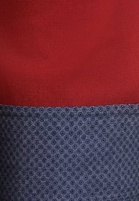 Rood en marineblauwe top met een textuurstof, waarbij de bovenste helft een effen rode kleur heeft en de onderste helft een cirkelpatroon op een donkere achtergrond toont.