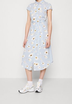 Robe bleu clair avec un motif floral, manches courtes, col et taille à nœud. Associée à des baskets blanches et des chaussettes de cheville.
