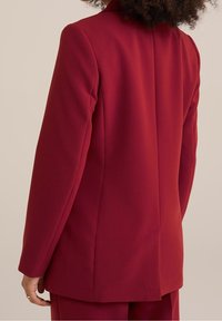 Czerwony żakiet o strukturalnym fasonie, z gładką fakturą tkaniny, długimi rękawami i dopasowanym krojem. Brak widocznych guzików lub ozdób.