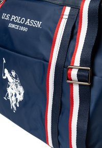 Borsa tote in nylon blu navy con accenti a righe bianche e rosse. Finiture dorate e logo di un giocatore di polo bianco ben visibile.