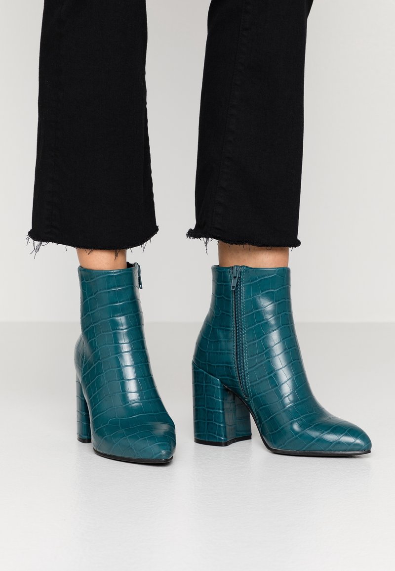 Bottines en cuir bleu sarcelle à motif crocodile, avec talons carrés et fermetures éclair latérales, portées avec un pantalon noir court sur fond gris.