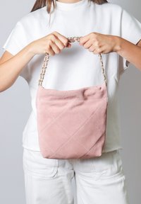 Borsa a tracolla in suede rosa con design trapuntato e motivi a diamante. Presenta una tracolla a catena. Tenuta da una persona in un outfit bianco su uno sfondo grigio.
