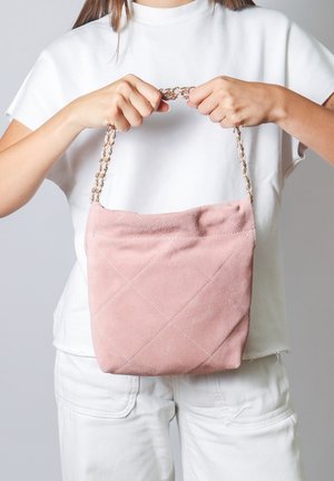 Roze suède handtas met een quilted ontwerp en diamanten patronen. Voorzien van een kettingriem. Vastgehouden door een persoon in een witte outfit tegen een grijze achtergrond.