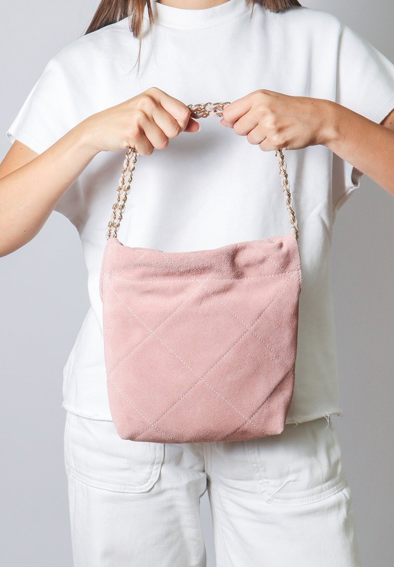 Borsa a tracolla in suede rosa con design trapuntato e motivi a diamante. Presenta una tracolla a catena. Tenuta da una persona in un outfit bianco su uno sfondo grigio.