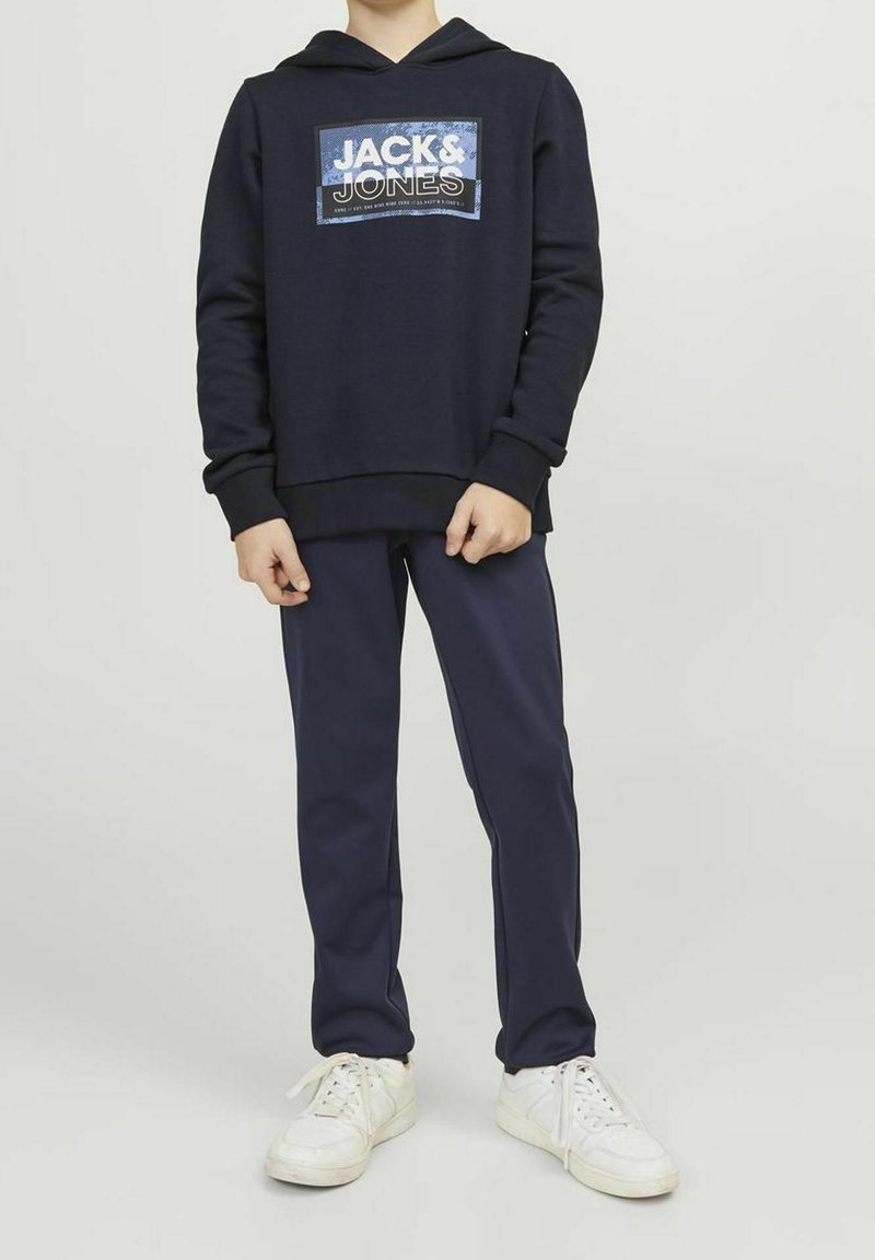 Hoodie bleu marine avec logo sur le devant, en matériau en coton, avec des poignets côtelés, associé à un pantalon bleu marine assorti et des baskets blanches. Design simple et décontracté.