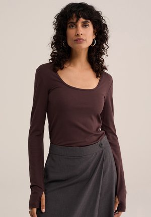 Femme aux cheveux bouclés et foncés porte un haut marron foncé à manches longues et une jupe portefeuille gris foncé, debout devant un fond beige uni.